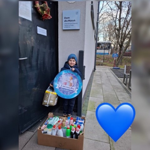 Chłopiec trzymający logo akcji "Paczuszka dla Maluszka" wraz ze skrzynią pełnych darów. Stoi przed Domem dla Samotnych Matek